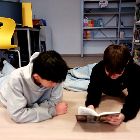 Zwei junge Jungen liegen auf einem Holzboden in einer Bibliothek. Einer liest ein Buch, während der andere ihn ansieht. Ein gelber Stuhl und ein Regal voller Bücher sind im Hintergrund zu sehen.