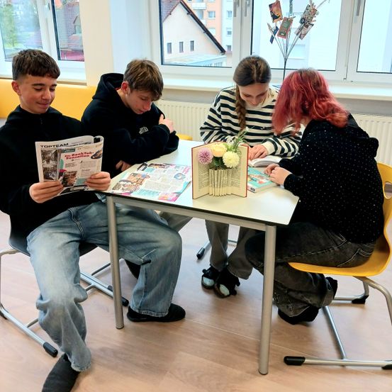 Vier Schüler sitzen um einen Tisch herum. Zwei lesen, einer schaut in ein Buch und einer schaut in eine Zeitschrift.