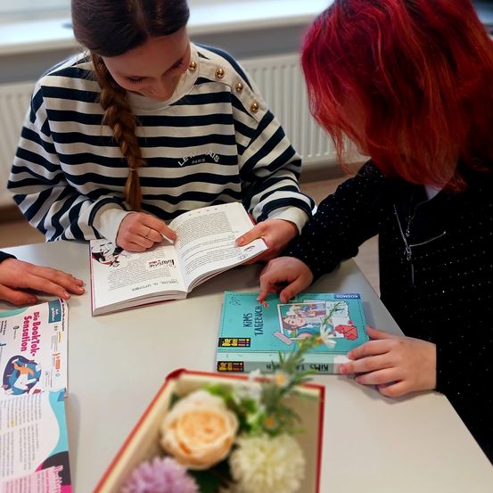 Zwei Mädchen lesen ein Buch an einem Tisch. Eine trägt ein gestreiftes Shirt und die andere hat rote Haare. Ein Blumenarrangement und andere Bücher sind auf dem Tisch.