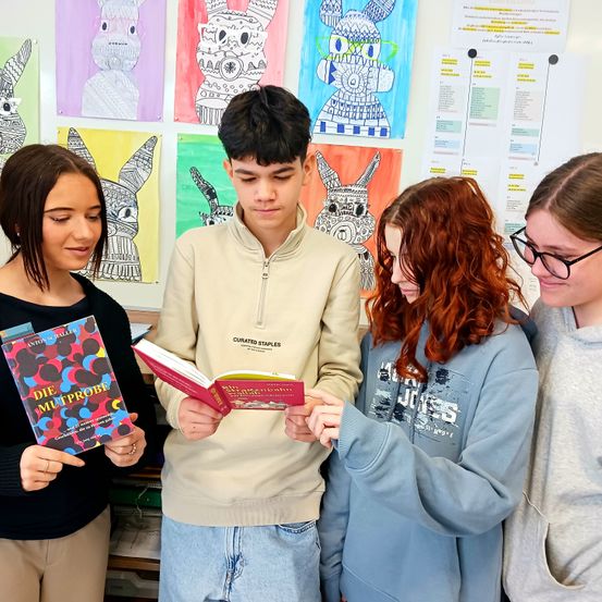 Vier junge Menschen in einem Klassenzimmer schauen auf ein Buch. Das Buch ist offen, und der Titel ist sichtbar. Hinter ihnen sind bunte Kunstwerke an der Wand angebracht.