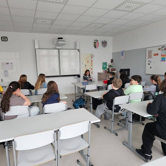 Ein Klassenzimmer mit Tischen und Stühlen in Reihen, eine Lehrerin liest vor. Der Raum hat weiße Wände und Decken, mit einer Tafel und einem Projektor. Die Schüler sitzen, einige schauen zur Lehrerin.