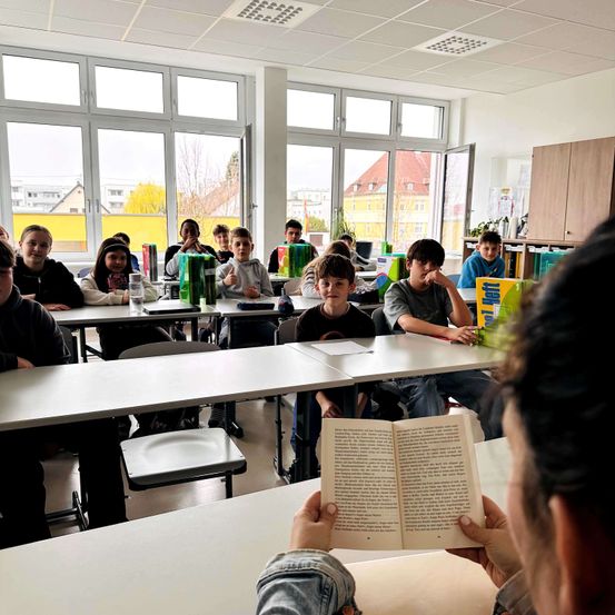 Ein Klassenzimmer mit Schülern an Tischen. Ein Schüler liest ein Buch, während andere zuhören. Der Raum hat große Fenster und eine helle Atmosphäre.