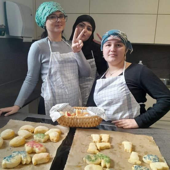Drei Frauen in Schürzen stehen in einer Küche. Eine macht ein Friedenszeichen. Ein Korb mit Keksen steht auf der Theke. Kekse sind auch auf Backblechen.