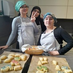 Drei Frauen in Schürzen stehen in einer Küche. Eine macht ein Friedenszeichen. Ein Korb mit Keksen steht auf der Theke. Kekse sind auch auf Backblechen.