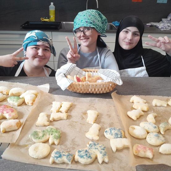 Drei Frauen posieren mit Friedenszeichen. Sie sind in einer Küche. Es gibt Kekse auf Backblechen. Ein Korb mit Keksen steht vor ihnen.
