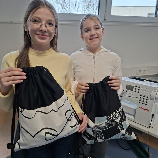 Zwei Mädchen lächeln und halten Taschen in einem Raum. Eine Tasche ist schwarz und weiß, die andere schwarz mit einem Aufnäher. Hinter ihnen steht eine Nähmaschine.