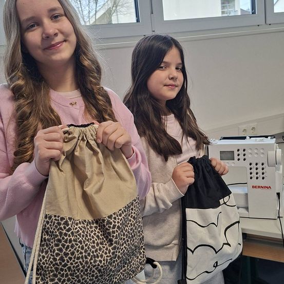 Zwei junge Mädchen stehen nebeneinander und halten Taschen. Eine hat eine Tasche mit Leopardenmuster und die andere hat eine schwarz-weiße Tasche. Sie sind in einem Raum mit einer Nähmaschine.