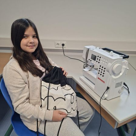 Ein junges Mädchen sitzt an einem Tisch mit einer Nähmaschine und hält eine Kordelzugtasche mit einem Cartoon-Charakter-Design. Sie lächelt und scheint sich in einem Klassenzimmer zu befinden.