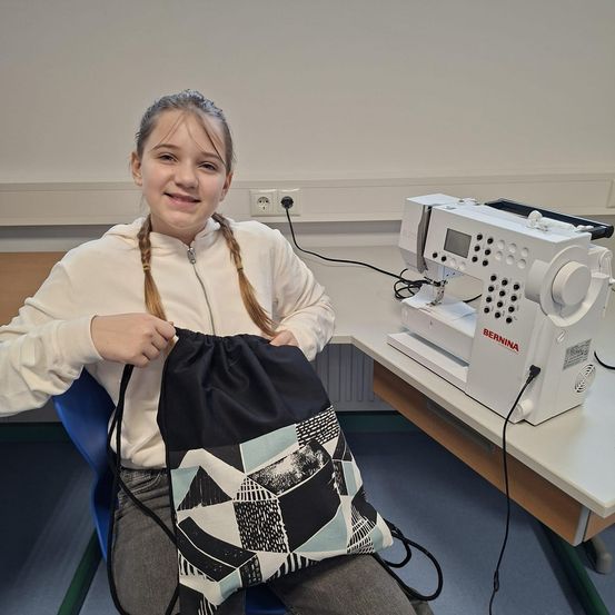 Ein junges Mädchen mit Zöpfen sitzt in einem blauen Stuhl im Klassenzimmer und hält eine Tasche mit geometrischem Muster. Hinter ihr steht eine Bernina-Nähmaschine auf einem Schreibtisch.