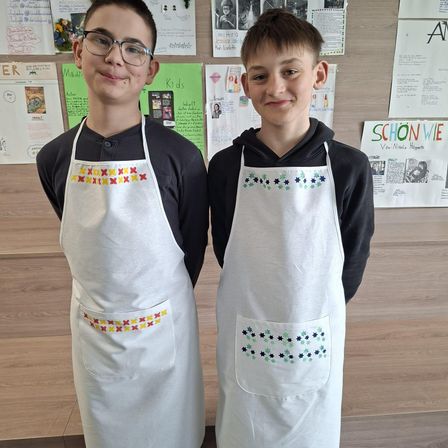 Zwei Jungen in weißen Schürzen mit gestickten Mustern, die in einem Klassenzimmer stehen. Der Junge auf der linken Seite hat eine Brille und ein kreuzförmiges Design.
