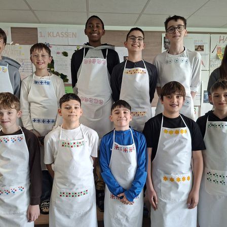 Eine Gruppe junger Jungen, die Schürzen mit verschiedenen Mustern tragen, posiert für ein Foto in einem Klassenzimmer. Das Schild hinter ihnen steht '1. Klassen'.
