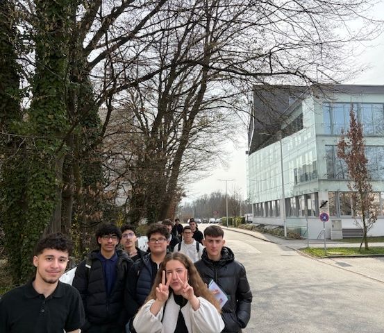 Eine Gruppe von Studenten in Winterkleidung steht an einer Straße, die von Bäumen gesäumt ist, und einem Gebäude mit Glasfenstern. Ein Student macht ein Friedenszeichen.