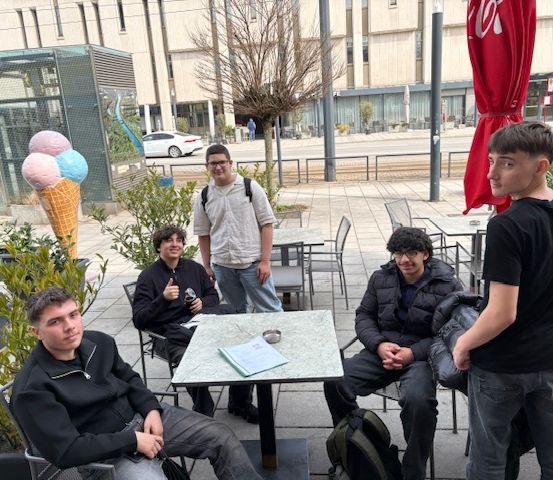 Fünf junge Männer sitzen auf Stühlen um einen Tisch in einem Café im Freien. Einer steht hinter ihnen. Sie sind in der Nähe von großen Eisbechern, einem roten Schirm und einem großen Gebäude.