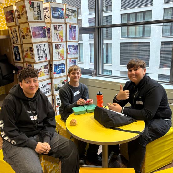 Drei Jungen sitzen an einem gelben Tisch in einem Raum mit großen Fenstern. Sie posieren für ein Foto. Ein Gebäude ist außerhalb der Fenster sichtbar.