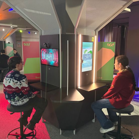 Zwei Personen sitzen auf Hockern vor einem interaktiven Display-Stand mit Bildschirmen und farbenfrohen Paneelen. Der Stand zeigt zwei Monitore mit farbenfrohen Grafiken.