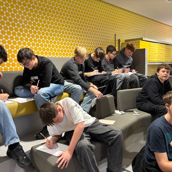 Eine Gruppe junger Jungen sitzt auf Bänken und schreibt mit Bleistiften auf Papier. Der Raum ist mit gelben Sechseckmuster-Wänden und gelben Schränken geschmückt.