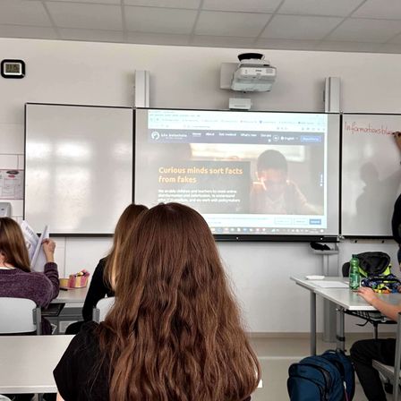 Ein Klassenzimmer mit einer Projektionsleinwand, die 'Neugierige Köpfe sortieren Fakten von Fakes' anzeigt. Schüler sitzen und sind in die Lektion vertieft.