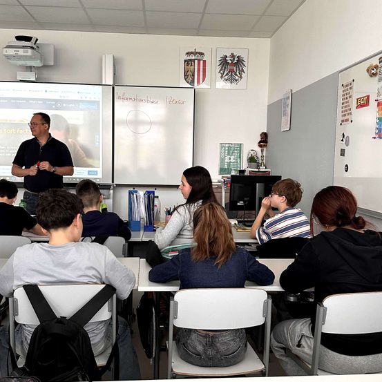 Ein Klassenzimmer mit Schülern, die aufmerksam einem Lehrer vorne lauschen, eine Projektionsleinwand mit einer Lektion und verschiedene Dekorationen und Poster an den Wänden.
