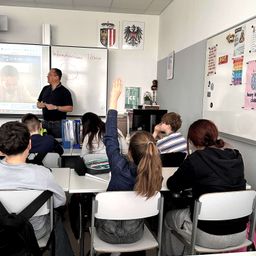 Ein Klassenzimmer mit Schülern, die aufmerksam einem Lehrer zuhören, der eine Präsentation auf einem Bildschirm hält. Der Lehrer trägt eine Brille und eine Uhr, und die Schüler sind engagiert.
