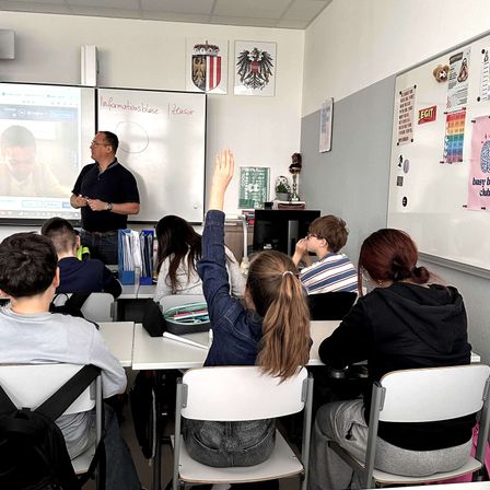 Ein Klassenzimmer mit Schülern, die aufmerksam einem Lehrer zuhören, der eine Präsentation auf einem Bildschirm hält. Der Lehrer trägt eine Brille und eine Uhr, und die Schüler sind engagiert.