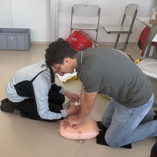 Zwei Personen üben CPR an einem Dummy in einem Raum mit Lagerboxen und Stühlen im Hintergrund.