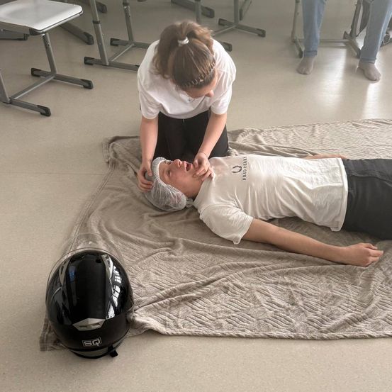 Eine Frau hilft einem Mann in einem weißen T-Shirt, der mit einem Helm in der Nähe auf einer Decke liegt. Sie sind in einem Klassenzimmer.