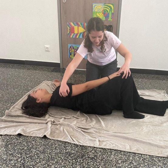 Eine Frau liegt auf einer Decke, während eine andere Frau über ihr steht und Druck auf ihren Rücken ausübt. Sie befinden sich in einem Raum mit einer Tür, die mit Gemälden verziert ist.