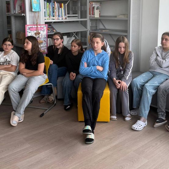 Eine Gruppe junger Mädchen sitzt auf einem gelben Hocker und Stühlen in einem Raum, vielleicht einem Klassenzimmer. Sie schauen mit verschränkten Armen etwas an.
