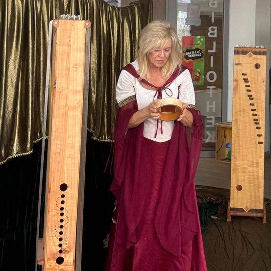 Eine Frau in historischer Kleidung steht mit einem hölzernen Instrument, möglicherweise einer Harfe, in den Händen. Sie schaut nach unten und scheint das Instrument zu untersuchen. Die Umgebung umfasst einen goldenen Vorhang und eine Glaswand.