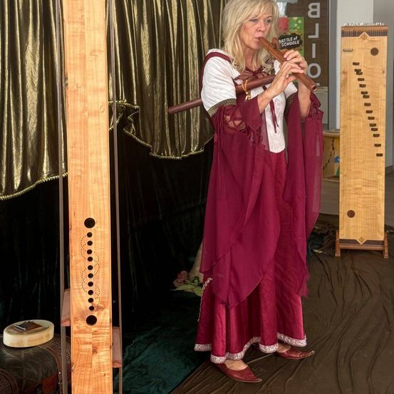 Eine Frau in einem burgunderroten Kleid und einem weißen Oberteil spielt eine Holzflöte. Sie steht in einem Raum mit einem braunen Vorhang und einer hölzern Harfe im Hintergrund.
