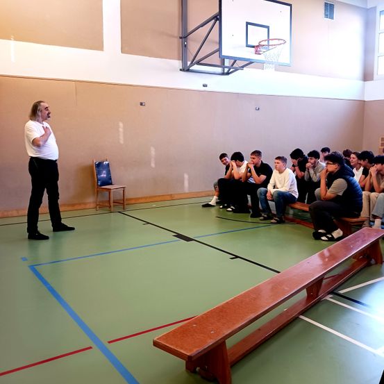 Ein Mann steht in einer Turnhalle und spricht zu Schülern, die auf Bänken sitzen. Der Raum hat Basketballkörbe, blaue und rote Linien auf dem Boden und einen Holzstuhl.