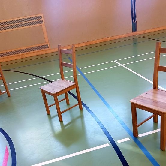 Zwei Holzstühle stehen auf einem Hallensportplatz, wahrscheinlich in einer Turnhalle. Der Platz hat grüne und blaue Markierungen und eine weiße Linie verläuft über den Boden.