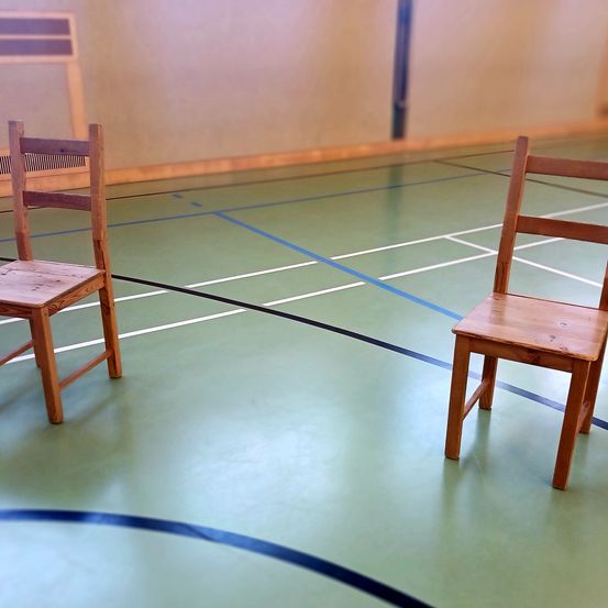 Zwei hölzerne Stühle stehen auf einem Basketballfeld mit blauen und weißen Linien. Der Boden ist grün und die Wände weiß.