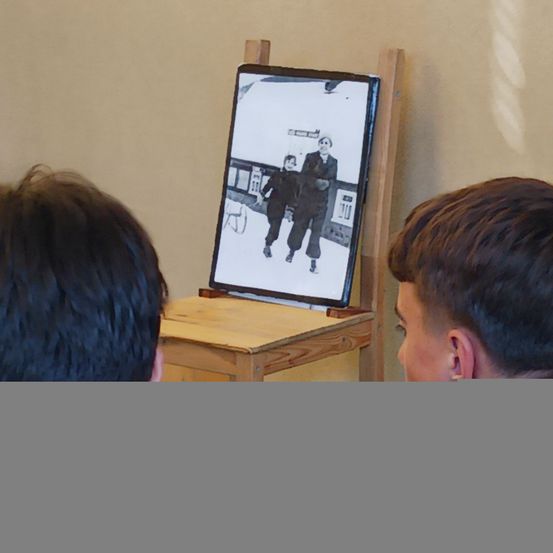 Zwei Jungen schauen sich ein Schwarz-Weiß-Foto auf einem kleinen Monitor an, der auf einem hölzernen Staffelei montiert ist. Das Foto zeigt zwei Frauen, die im Schnee gehen.