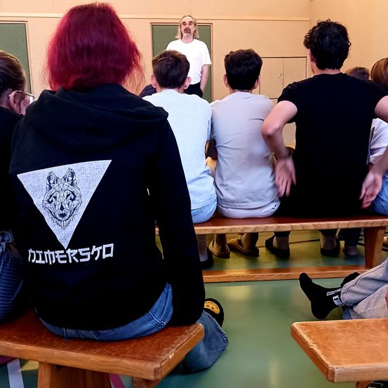 Eine Gruppe von Schülern in Freizeitkleidung sitzt in einem Raum und beobachtet einen Mann in einem weißen Hemd vorne. Der Raum hat einen Holzboden und eine Wand mit einer Tafel.
