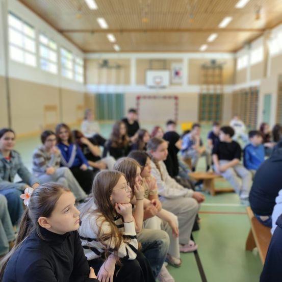 Eine Gruppe von Kindern und Jugendlichen sitzt auf Bänken in einer Turnhalle, einige schauen nach vorne, während andere eine Präsentation verfolgen.