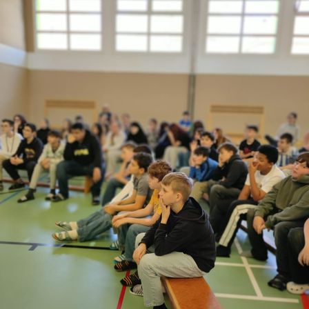 Eine Gruppe junger Schüler sitzt auf Bänken in einer Turnhalle und hört einem Sprecher zu. Sie sind aufmerksam und engagiert.