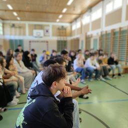 In einer Sporthalle sitzt eine Gruppe von Schülern in Reihen und hört aufmerksam einem Redner zu. Der Basketballplatz und die Fenster sind im Hintergrund zu sehen.
