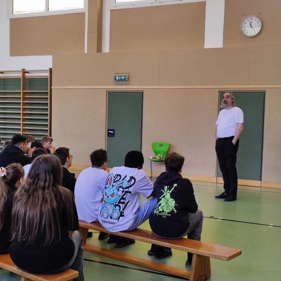 Ein Mann steht vor einer sitzenden Gruppe in einem Raum, trägt ein weißes T-Shirt und schwarze Hose. Der Raum hat einen grünen Boden und eine Uhr an der Wand.