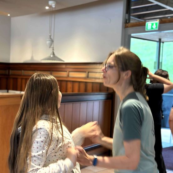 Zwei Frauen tanzen in einem Innenbereich mit Holzwänden. Eine Frau trägt eine Uhr und eine Brille, und die andere hat langes Haar. Dahinter steht eine Person in der Nähe des Ausgangs.