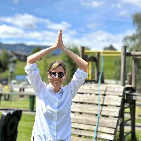 Eine Frau in einem weißen Hemd und Sonnenbrille praktiziert Yoga in einem Park mit einem Spielplatz im Hintergrund.