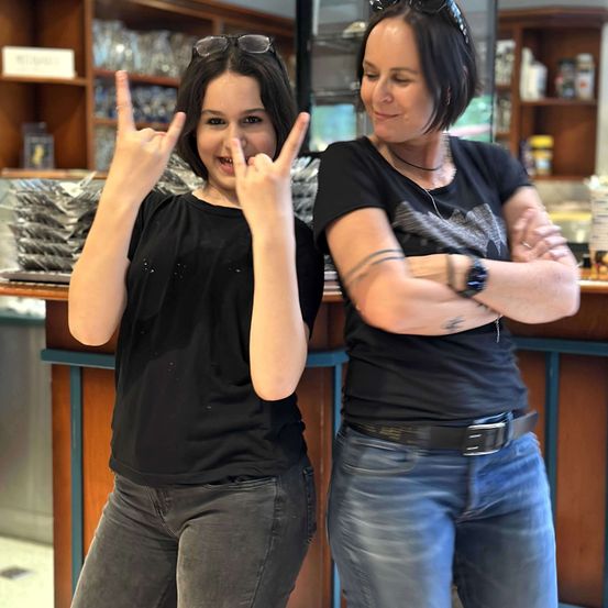 Zwei Frauen in schwarzen T-Shirts, eine mit Sonnenbrille auf dem Kopf und die andere mit einer Uhr, stehen in einer Küche.