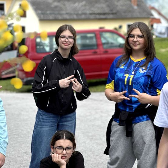 Drei Frauen posieren für ein Foto. Zwei tragen Brillen, eine in einer schwarzen Jacke, eine andere in einem blau-gelben Shirt. Ein roter Van steht im Hintergrund.