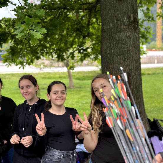 Drei Mädchen machen Friedenszeichen mit ihren Händen. Sie stehen unter einem Baum, mit vielen Pfeilen am Stamm. Sie tragen schwarze T-Shirts.