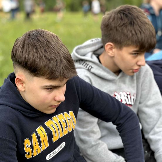 Zwei junge Jungen sitzen auf Gras, einer mit einer San Diego-Hoodie. Der Junge auf der rechten Seite trägt eine graue Hoodie mit Text. Verschwommene Figuren sind im Hintergrund.