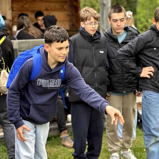 Eine Gruppe von Jungen steht auf einem Feld mit einer hölzernen Struktur im Hintergrund. Ein Junge mit einem Rucksack zeigt auf einen anderen.