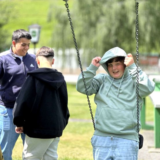 Drei Jungen sind im Freien. Ein Junge ist auf einer Schaukel und lächelt. Ein anderer Junge schaut ihn an, während der dritte Junge hinter ihm steht und lächelt. Dahinter sind ein Laternenpfahl und eine Bank zu sehen.