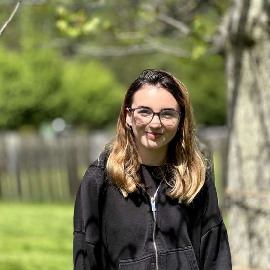 Eine Frau mit blonden Haaren, die eine Brille und eine schwarze Kapuzenjacke trägt, steht in einem Park mit einem Baum und einem Zaun hinter ihr.