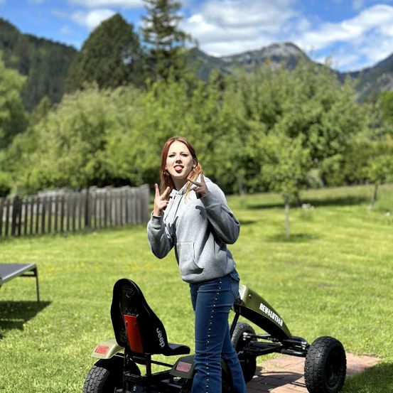 Eine Frau in einer grauen Kapuzenjacke und Jeans steht neben einem grünen Go-Kart auf einem Rasenfeld mit einem Holzzaun und Bäumen im Hintergrund.