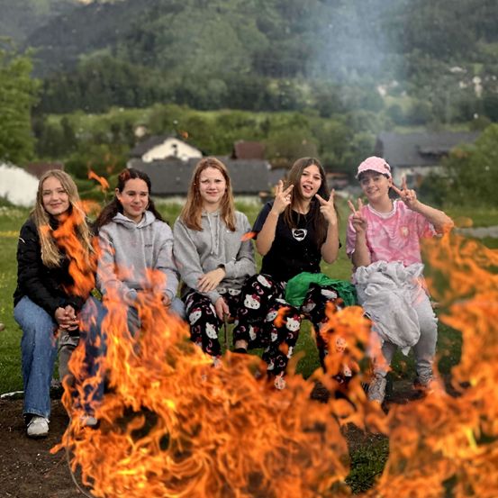 Fünf junge Frauen sitzen um ein Lagerfeuer und lächeln alle mit Friedenszeichen. Sie sind von einer grünen Landschaft und Bergen im Hintergrund umgeben.